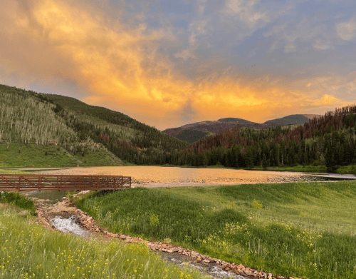 A serene landscape featuring a lake at sunset, surrounded by mountains and lush green fields.