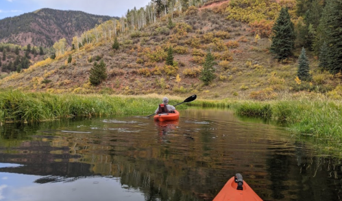 A person kayaking on a calm river surrounded by lush greenery and autumn-colored hills.