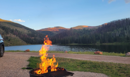 A campfire burns in a grill by a serene lake, surrounded by mountains and trees under a clear blue sky.