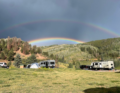 A scenic campsite with RVs and tents under a double rainbow and dark storm clouds over green mountains.