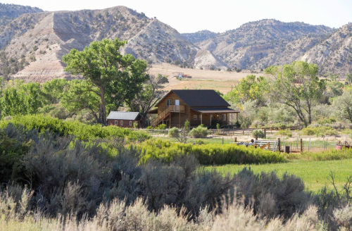 A rustic house surrounded by greenery and mountains under a clear blue sky.