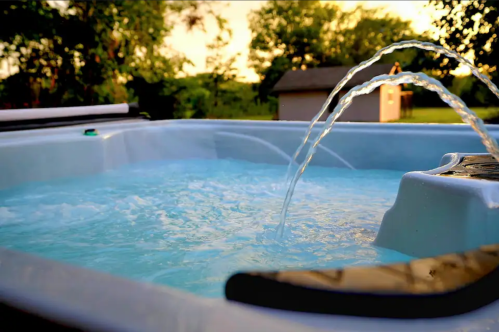 A hot tub with bubbling water and a fountain feature, surrounded by greenery and a sunset sky in the background.