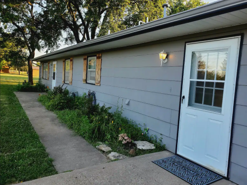A gray house with a white door, surrounded by greenery and a concrete pathway leading to the entrance.