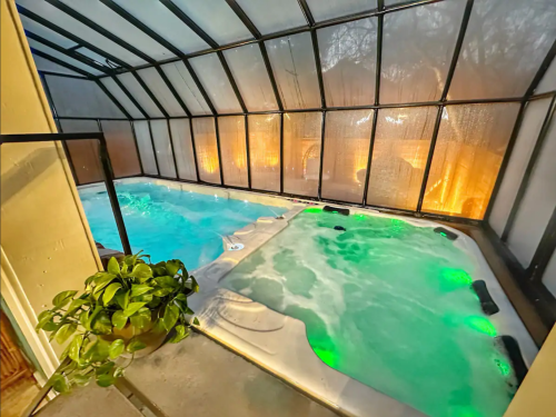 Indoor pool and hot tub with green lights, surrounded by glass walls and a potted plant in the corner.