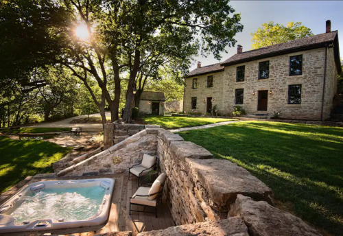 A serene outdoor scene featuring a hot tub, stone wall, and a historic house surrounded by trees and lush greenery.