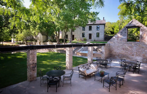 A serene outdoor space with stone ruins, green trees, and seating areas, overlooking a historic building.