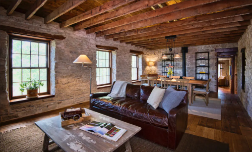 Cozy living room with stone walls, wooden beams, a leather sofa, and a dining area with a table and chairs.