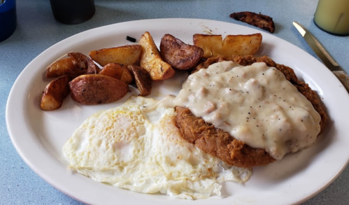 A plate of fried chicken topped with gravy, two fried eggs, and seasoned potato wedges.