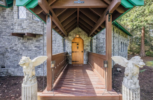 A wooden entrance with a stone archway, flanked by decorative pillars and angel statues, leading to a rustic door.