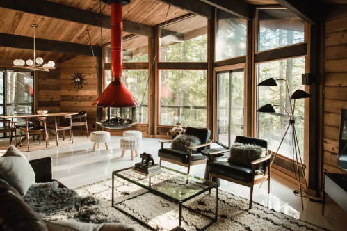 Cozy modern living room with large windows, a red fireplace, and stylish furniture, surrounded by trees.