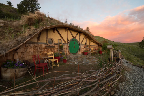 A charming hobbit-style home with a round green door, surrounded by colorful flowers and rustic furniture at sunset.