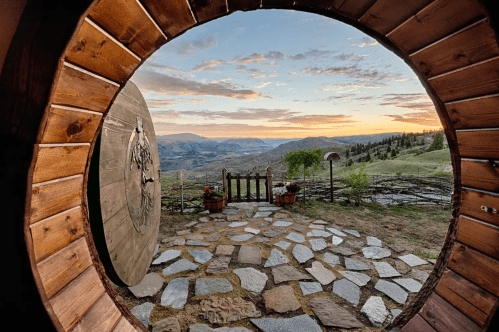 View through a round wooden door, showcasing a scenic landscape at sunset with stone pathway and flower pots.