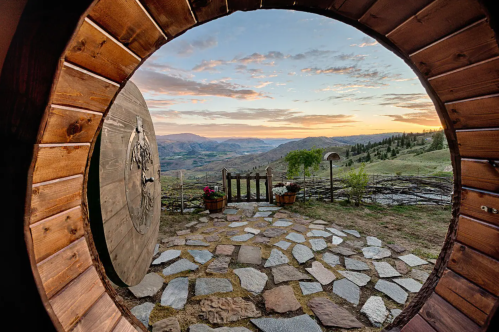 View through a round wooden door, showcasing a scenic landscape at sunset with stone pathway and flower pots.