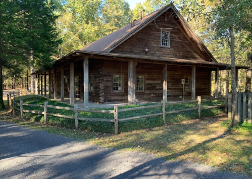 A rustic wooden house surrounded by trees, with a gravel path and a wooden fence in the foreground.
