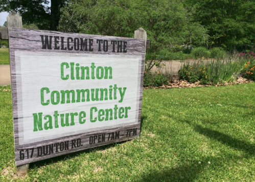Sign welcoming visitors to the Clinton Community Nature Center, with location and hours listed. Green grass and plants in background.