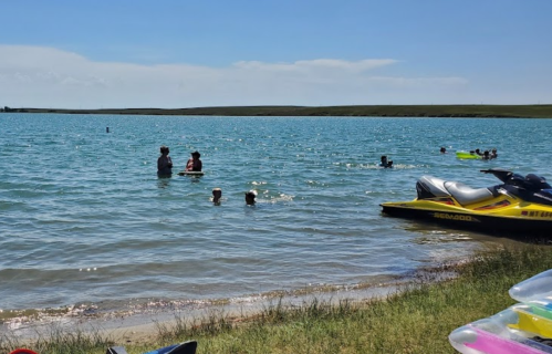 People swimming and playing in a blue lake, with jet skis parked on the shore under a clear sky.