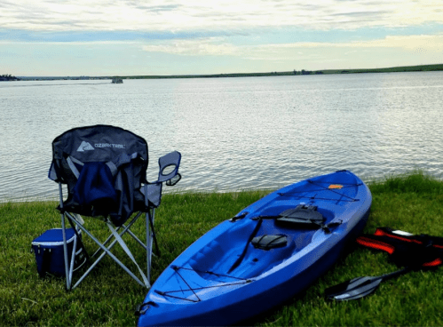 A blue kayak and a camping chair sit on green grass by a calm lake under a cloudy sky.