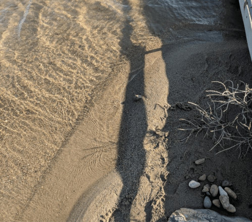 A sandy shore with gentle waves, pebbles, and a shadow cast by a person standing near the water's edge.