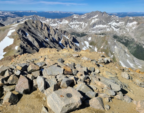 Rocky mountain summit with snow-capped peaks in the distance under a clear blue sky.