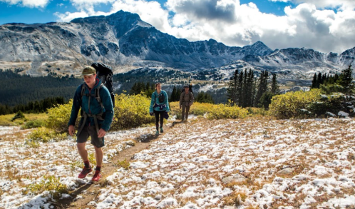 Three hikers trek along a snowy trail with mountains in the background under a partly cloudy sky.