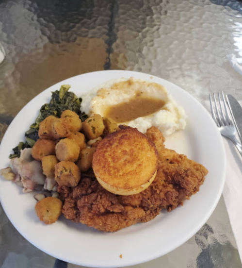A plate of fried chicken with a biscuit, collard greens, okra, mashed potatoes, and gravy on a glass table.