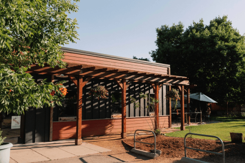 A modern building with wooden pergolas, surrounded by greenery and outdoor seating under a clear blue sky.