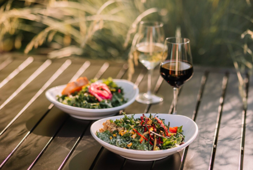 Two bowls of fresh salads on a table, accompanied by glasses of white and red wine, with greenery in the background.