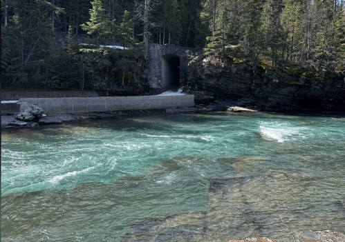 A turquoise river flows past rocky banks and a stone tunnel, surrounded by lush green trees.