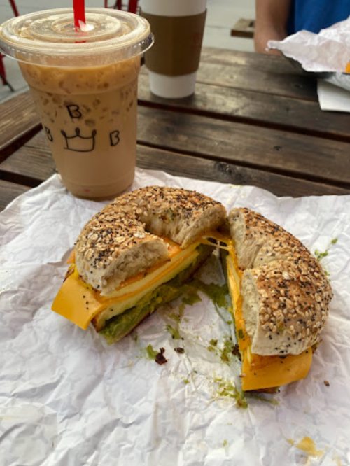 A toasted bagel sandwich with cheese and avocado, alongside an iced coffee on a wooden table.