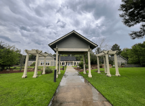 A pathway leads to a gray building under a cloudy sky, framed by white pergolas and green grass on either side.
