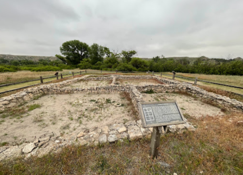 Ruins of a stone structure with a historical sign, surrounded by grass and trees under a cloudy sky.