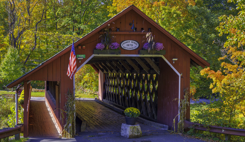 A red covered bridge surrounded by colorful autumn foliage, with flowers and an American flag at the entrance.