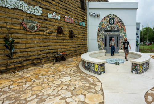 Two people walk past a colorful, mosaic-adorned entrance to a building, surrounded by a stone pathway.