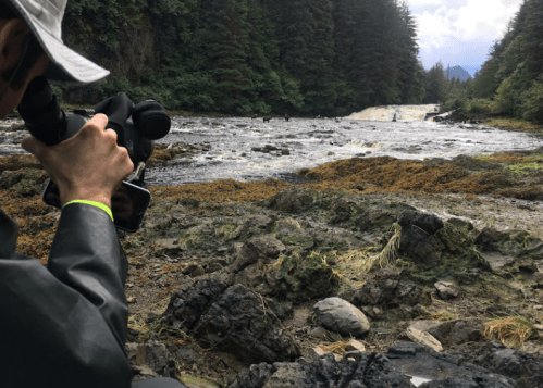 A person in a hat photographs a river with a waterfall, surrounded by lush greenery and rocky terrain.