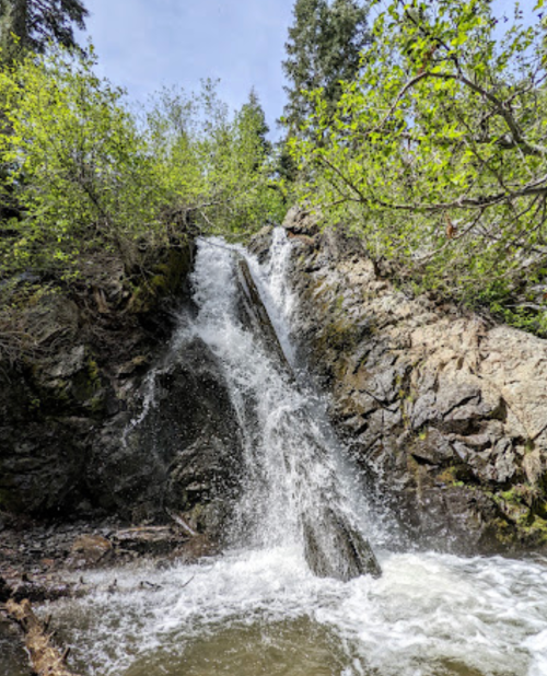 A cascading waterfall flows over rocky terrain, surrounded by lush green trees and a clear blue sky.