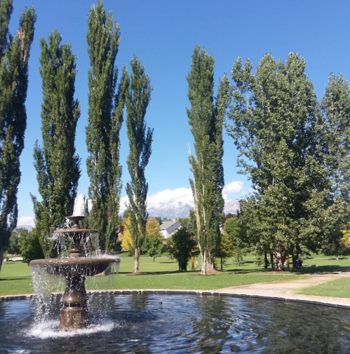A fountain in a park surrounded by tall trees and a clear blue sky, with mountains in the background.