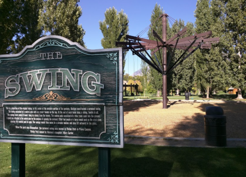 A playground swing set with a large sign titled "The Swing" in a park surrounded by trees and gravel.