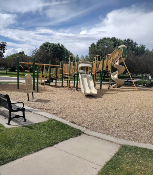 A playground with slides and climbing structures, surrounded by gravel and grass, under a partly cloudy sky.
