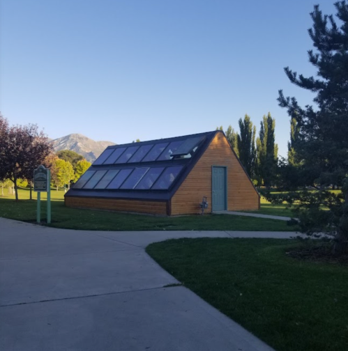 A modern wooden greenhouse with solar panels, surrounded by grass and trees, set against a mountain backdrop.