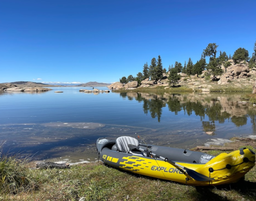 A yellow kayak rests on the shore of a calm lake, surrounded by trees and rocky terrain under a clear blue sky.