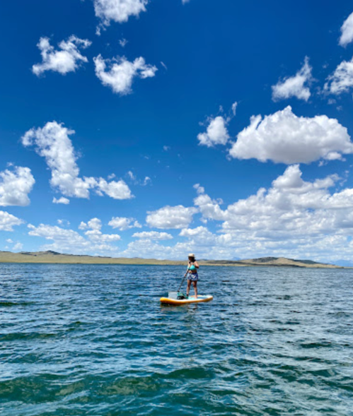 A person paddleboarding on a calm lake under a bright blue sky with fluffy white clouds.