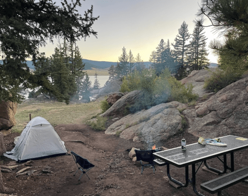 A serene campsite by a lake, featuring a tent, picnic table, and a small fire surrounded by trees and rocky terrain.