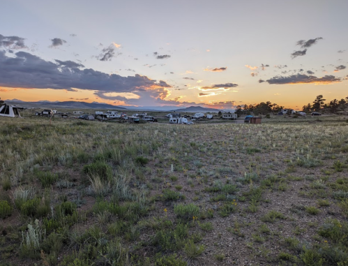 A scenic sunset over a grassy field with RVs and tents in the distance, surrounded by mountains and clouds.