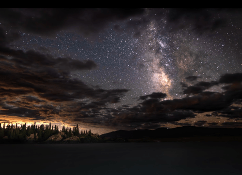 A starry night sky with the Milky Way, clouds, and a silhouette of trees and mountains reflected in calm water.