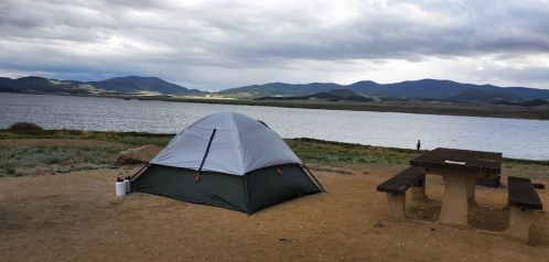 A tent set up by a lake, with mountains in the background and a cloudy sky overhead.