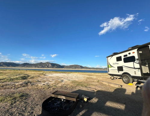 A camper parked by a lake with mountains in the background under a clear blue sky.