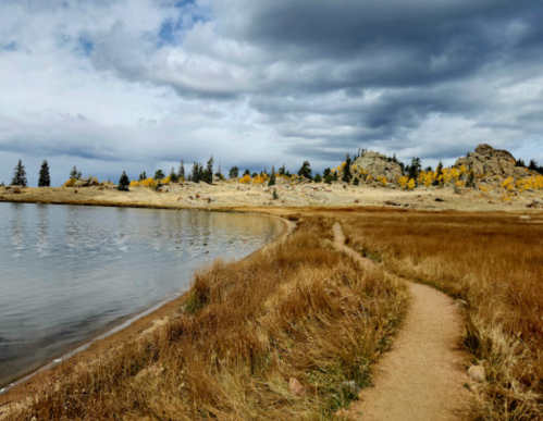 A winding path leads along a lake, surrounded by golden grass and rocky hills under a cloudy sky.