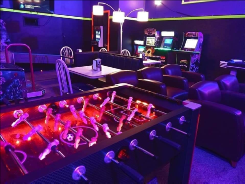 A vibrant game room featuring a foosball table, arcade machines, and cozy seating under colorful lighting.