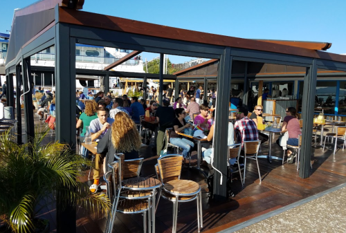 A bustling outdoor restaurant with many patrons enjoying food and drinks under a clear blue sky.