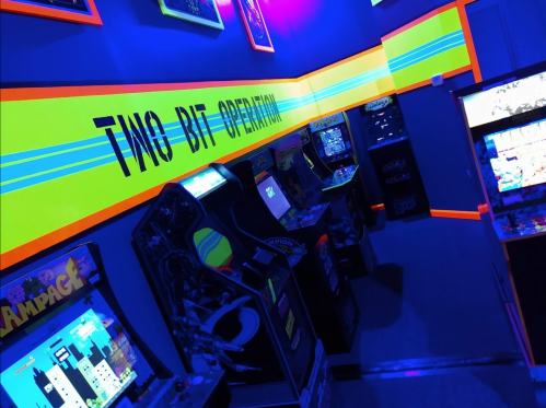 A vibrant arcade room with classic game machines and neon accents, featuring the text "TWO BIG OPERATION" on the wall.